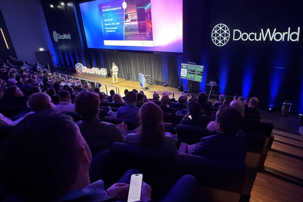 A large crowd is sitting in front of a stage with a large screen where live captions are shown